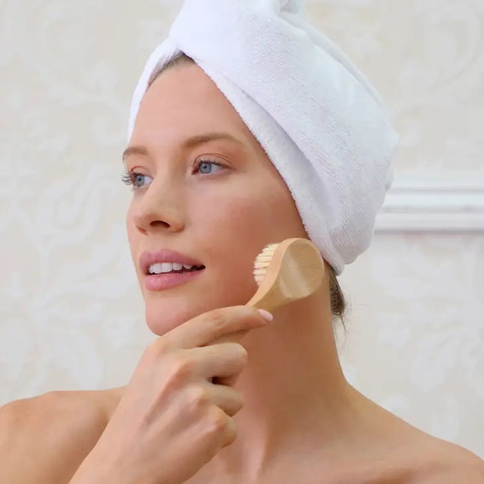 A DAILY CONCEPTS FACIAL DRY BRUSH with a wooden handle and natural bristles is displayed in front of its packaging, labeled for gentle exfoliation and radiant skin. The package shows product details and icons.