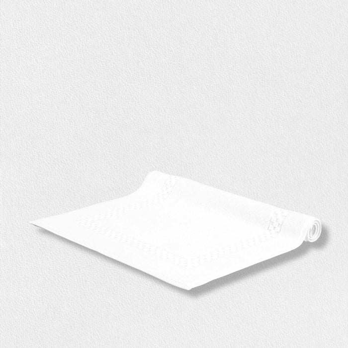 The FEARRINGTON LIFESTYLE TOWEL COLLECTION - BATH MATT by Frette North America lies partly unrolled on a light, textured surface, its elegant border and subtle design evoking refined luxury against a minimal monochromatic background.