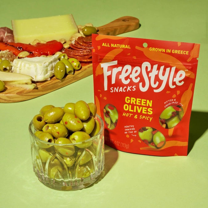 A red package of FREESTYLE - HOT & SPICY GREEN OLIVES by FREESTYLE SNACKS features Calabrian chili-accented green olives. The design highlights olives, chilies, "All Natural," "Grown in Greece," and emphasizes "Healthy Snacking on the Go." Net weight: 4 oz (113g).