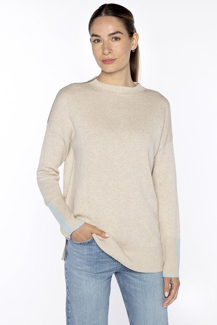 A woman stands against a white background in the KINROSS CASHMERE Doubleknit Lurex Trim Funnel Sweater in Dune and light blue jeans, her long hair pulled back as she smiles at the camera—an effortless travel look.