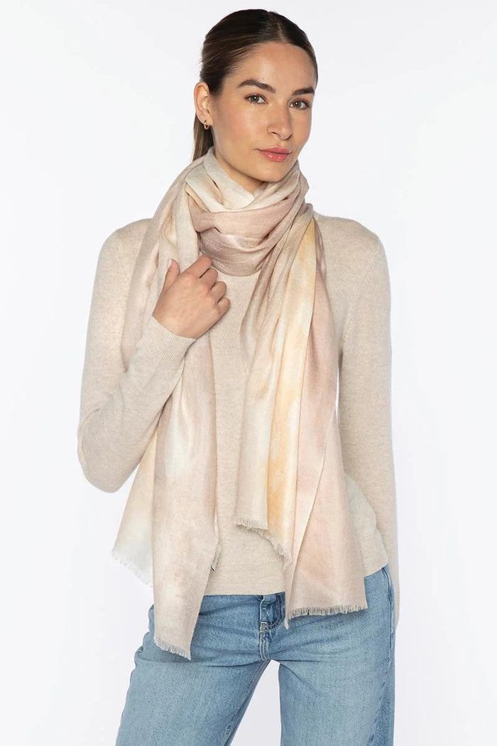 A woman in a light beige sweater and blue jeans poses against a white background, wearing the KINROSS CASHMERE - WATERCOLOR DOT SCARF IN CAMEO MULTI by KINROSS wrapped stylishly around her neck.