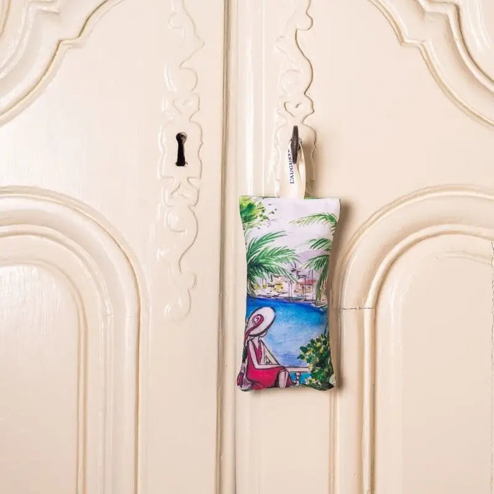 The L'AUGUSTE PROVENCE - FRENCH LAVENDER SACHET FROM PROVENCE is a small, handcrafted sachet featuring a hat-wearing woman by the water with palm trees, filled with organic lavender and labeled “L'AUGUSTE PROVENCE” for lasting freshness.