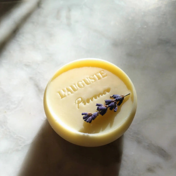 L'AUGUSTE PROVENCE - THE ESSENCE OF PROVENCE LAVENDER SOAP is a round, handmade French soap wrapped in painted landscape fabric, presented in a Provence gift box. Dried lavender rests nearby on a white cloth background.
