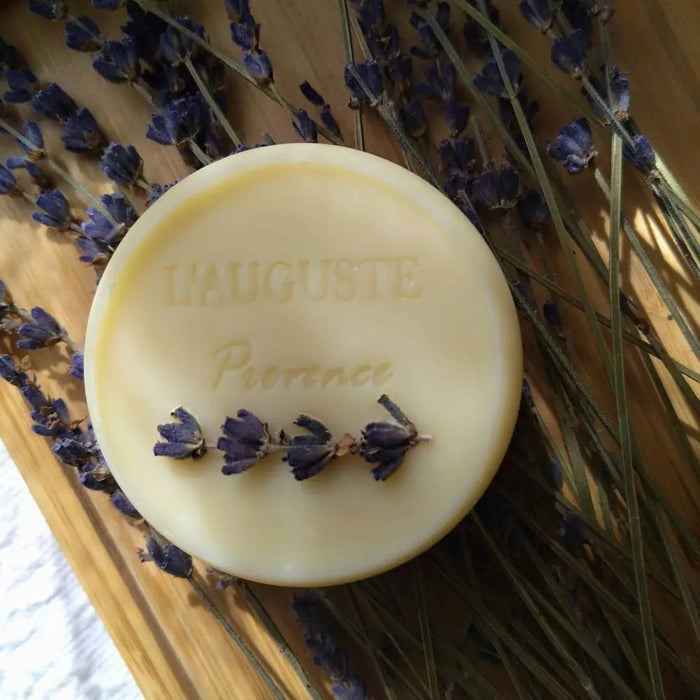 L'AUGUSTE PROVENCE – THE ESSENCE OF PROVENCE Lavender Soap, a round handmade French soap wrapped in cloth with a lavender field and tree motif, rests in an open L'AUGUSTE PROVENCE gift box beside its lid and scattered lavender buds.