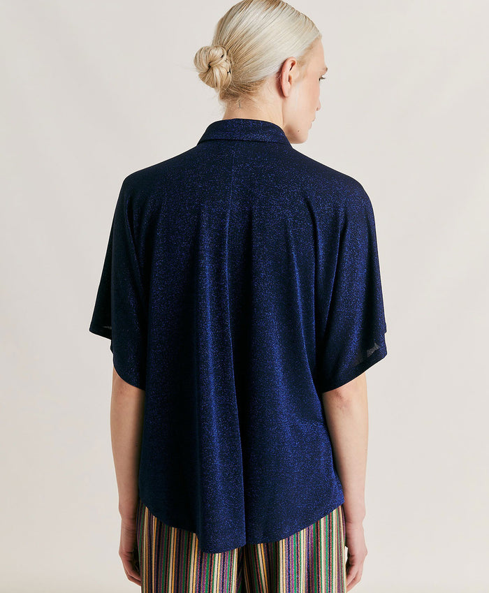 A person in a MOMONI - BROOKLYN blue shirt with a classic collar, paired with colorful beaded necklaces and tucked into striped pants. The loose-fitting, short-sleeve button-up stands out against the plain light background.