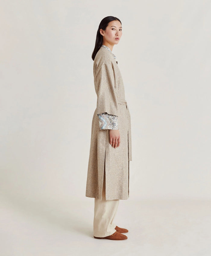 A woman poses against a plain light background wearing the MOMONI - CALL Lightweight Coat in Lurex Basketweave over a patterned shirt and loose, high-waisted beige wide-leg pants. She has straight dark hair and brown shoes.