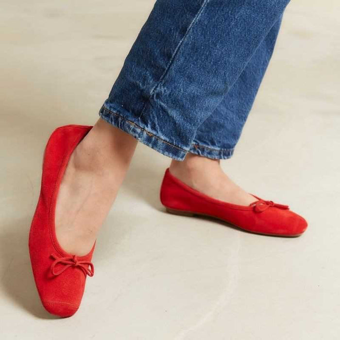 A person in white jeans, a beige coat, and carrying a round straw bag stands on light stone pavement, wearing REQINS HARMONY suede ballerina flats in rouge by REQINS.