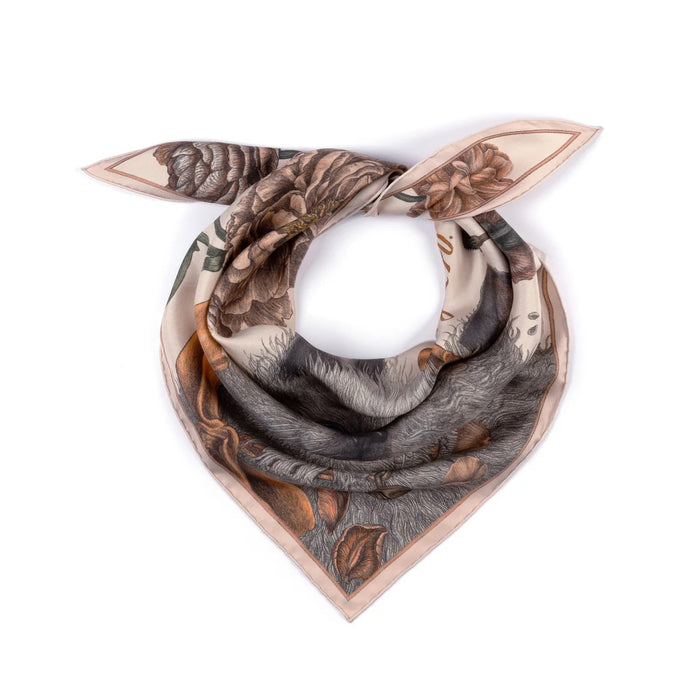 A person in a cream-colored shirt and skirt is seen from the back, wearing a SABINA SAVAGE - RING A RING O' ROSES silk twill triangle scarf by SABINA SAVAGE ENGLAND, featuring a fluffy Ragdoll kitten with blue eyes, framed by English garden flowers.