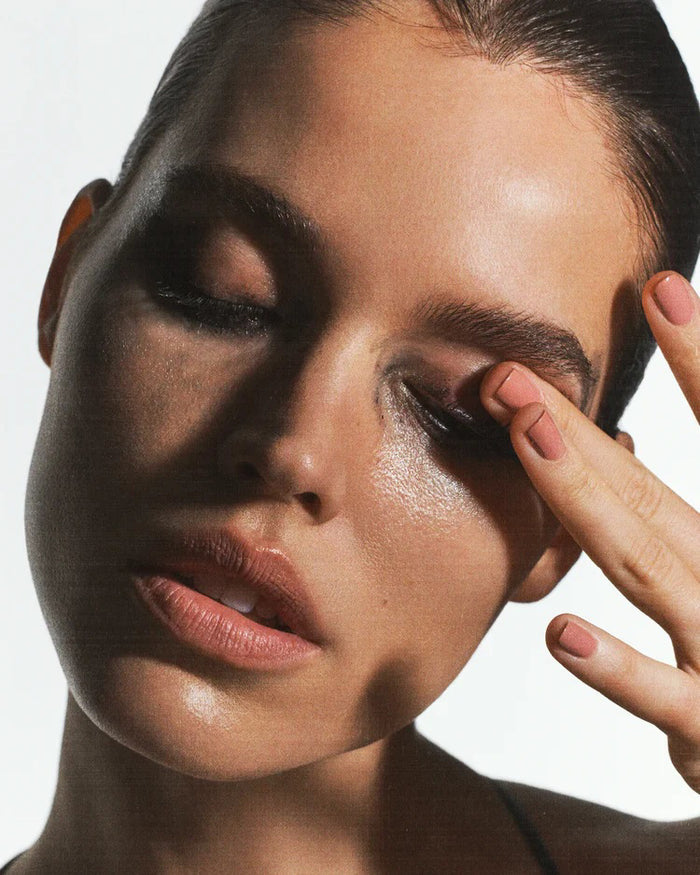 A woman with slicked-back hair touches her eyelid, smudging dark makeup—perhaps after using U BEAUTY's THE MANTLE SKIN CONDITIONING WASH 150ML. Her dewy skin and parted lips create a fresh, natural look as she tilts her head forward.