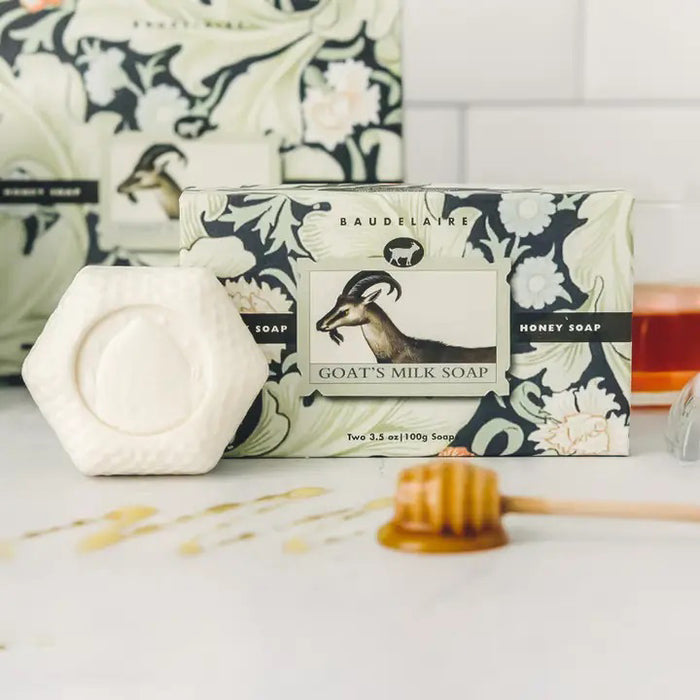 The BAUDELAIRE - GOATS MILK HONEY SOAP HEX BOX 3.5OZ comes in a hexagonal box adorned with a delicate floral motif and features a central label illustrated with a goat. Gentle enough for sensitive skin, this moisturizing soap is visible through an opening in the packaging.