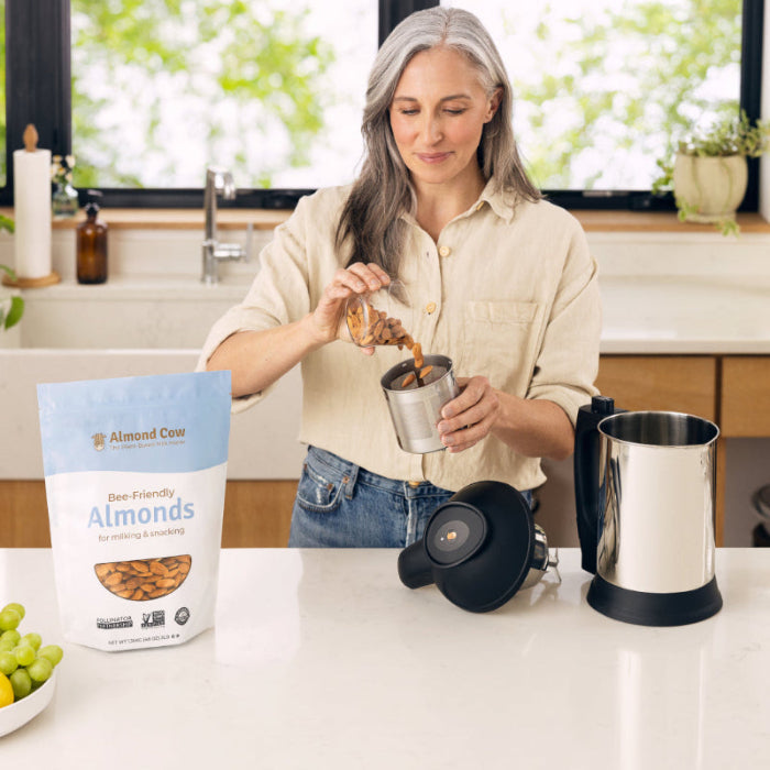 A middle-aged woman pours bee-friendly almonds into a milk maker machine, with a 3lb bag of organic almonds sitting nearby.