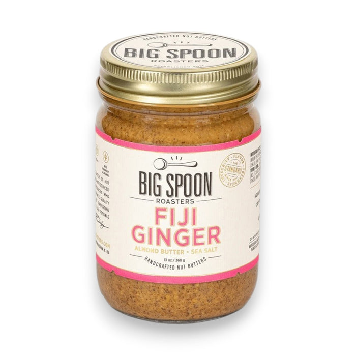 A jar of BIG SPOON ROASTERS - FIJI GINGER ALMOND, featuring a gold lid and a white label with pink accents that displays the brand name, flavor, and additional product information. The crystallized ginger-infused almond butter is visible through the clear jar.