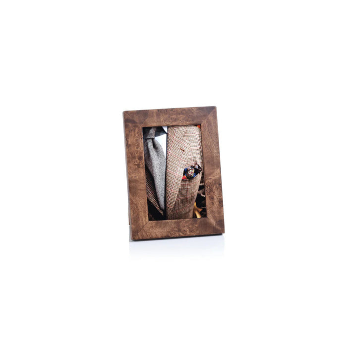 A gold table lamp stands beside the ZODAX BURL WOOD PHOTO FRAME 4X6 displaying a photo of a man in a suit, both set atop large books—including "Luxury Trains"—on a wooden surface with a gray backdrop.