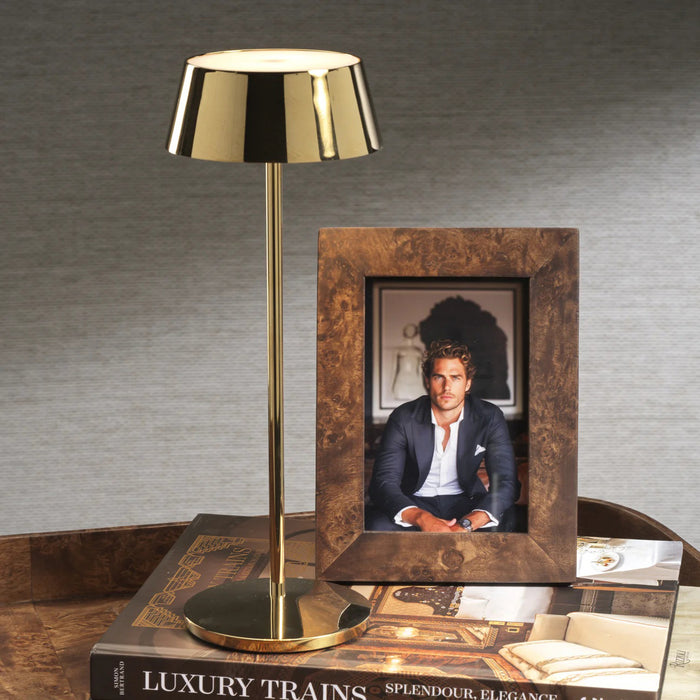 A gold table lamp stands beside the ZODAX BURL WOOD PHOTO FRAME 4X6 displaying a photo of a man in a suit, both set atop large books—including "Luxury Trains"—on a wooden surface with a gray backdrop.