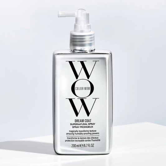 A bottle of KT SUPPLY COLORWOW - DREAM COAT SUPERNATURAL SPRAY, featuring a pump dispenser and black and white text on a shiny silver background, is placed on a light surface with soft shadowing.
