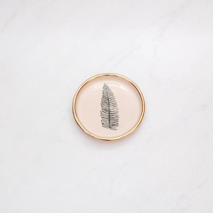 The COMPOST DESIGN - FERN TINY DISH 3" features a black fern leaf on white stoneware, highlighted by a 22k gold rim. Its light, neutral tones and hand-built rustic look beautifully contrast against the white marble surface.