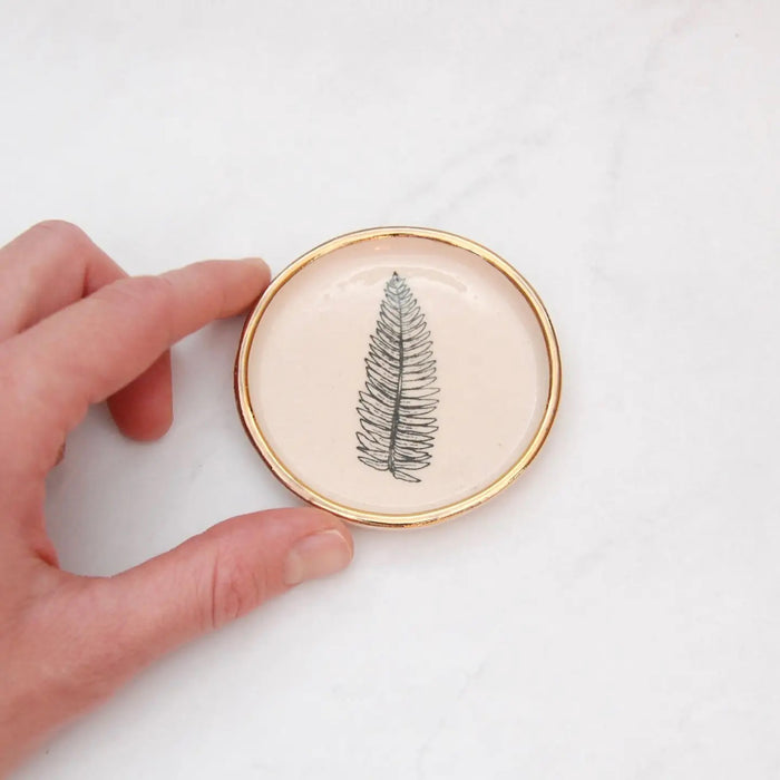 The COMPOST DESIGN - FERN TINY DISH 3" features a black fern leaf on white stoneware, highlighted by a 22k gold rim. Its light, neutral tones and hand-built rustic look beautifully contrast against the white marble surface.