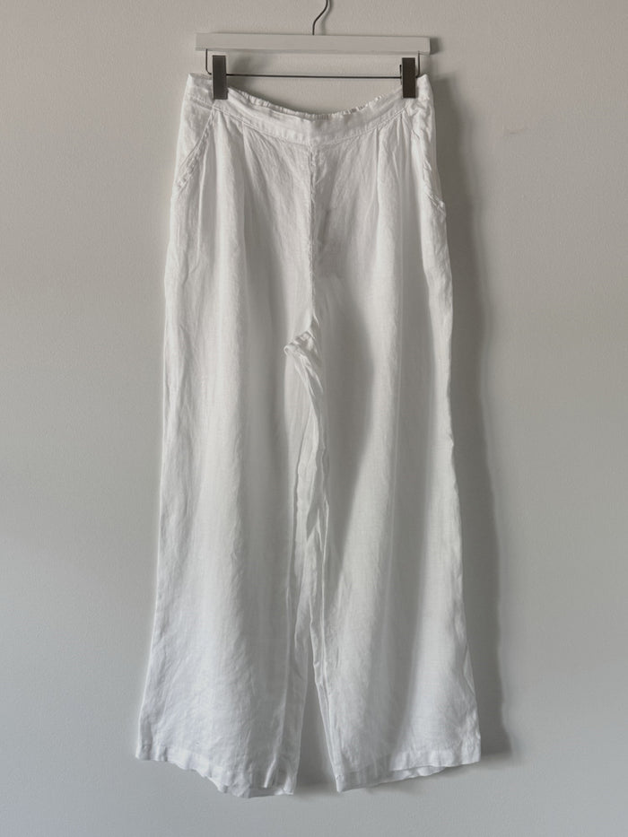 The CP SHADES - TESS LINEN WIDE LEG PANT by CP SHADES hangs on a black hanger against a light gray wall. These linen pants offer a relaxed fit with pockets, visible seams, and a slightly wrinkled texture.
