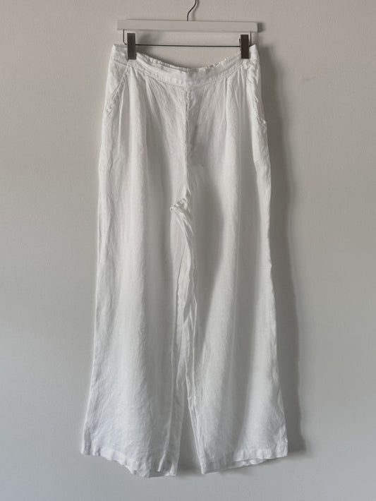 The CP SHADES - TESS LINEN WIDE LEG PANT by CP SHADES hangs on a black hanger against a light gray wall. These linen pants offer a relaxed fit with pockets, visible seams, and a slightly wrinkled texture.