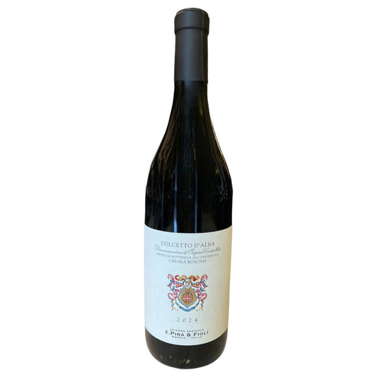 A bottle of E. PIRA DOLCETTO D'ALBA 2024 by WINE: GRAPEVINE, featuring a white label with a decorative crest and text, sits on a light-colored surface with blurred shelves in the background.