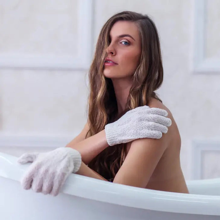 A package of DAILY CONCEPTS EXFOLIATING GLOVES appears beside a pair of white textured spa gloves. The white and purple packaging highlights their effectiveness for removing dead skin.