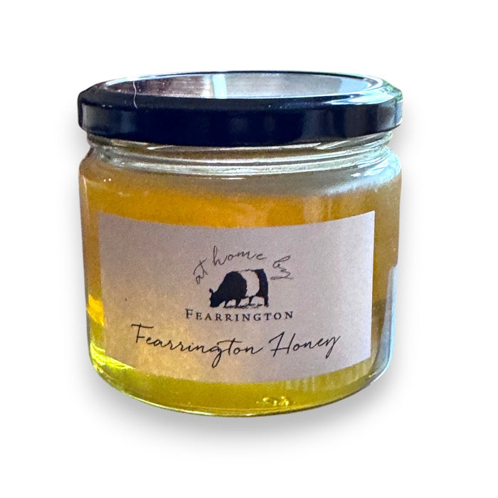 The Fearrington Signature Collection's hexagonal glass jar elegantly holds their Wildflower Honey, known for its raw quality. The label states: "Raw, Unfiltered. Handmade with care in our kitchens. May contain allergens. 282 grams, 10 oz." Inside is the finest golden honey from Fearrington House.