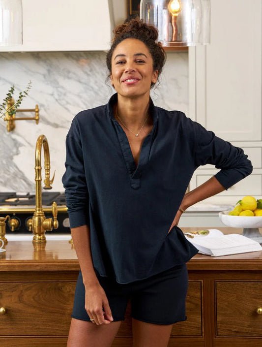 A woman wearing the FRANK & EILEEN Patrick Popover Henley in Vintage Jersey British Royal Navy and shorts stands smiling in a modern kitchen with brass fixtures, a marble backsplash, and a bowl of lemons on the counter.