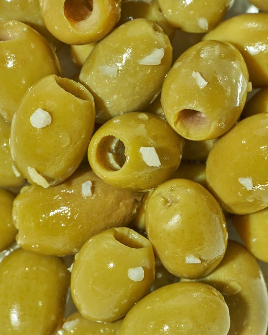 A yellow packet of FREESTYLE - LEMON GARLIC GREEN OLIVES from FREESTYLE SNACKS, featuring green olives flavored with extra virgin olive oil. The packaging highlights the "all-natural" ingredients and notes that the olives are "grown in Greece." Weighing 4 oz (113g), the pack showcases images of green olives alongside phrases such as "pitted & unpasteurized" and "healthy snacking on the go.