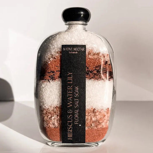 A glass bottle filled with layered bath salts in shades of pink, white, and brown. The black label reads "NATIVE NECTAR" at the top and "HIBISCUS & WATERLILY FLORAL SALT SOAK" vertically down the side, showcasing its mineral-rich sea salt properties. The bottle is set against a plain background.