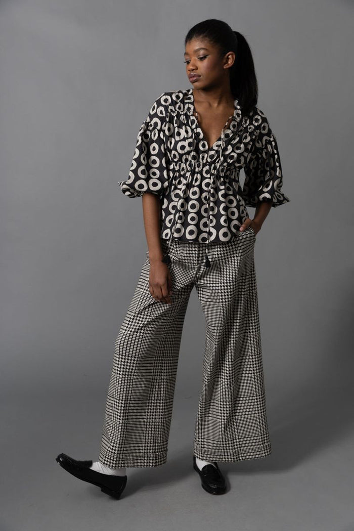 A person with long brown hair, seen from behind, wears the HOPE FOR FLOWERS Shirred Blouse and loose, dark green cargo pants with large pockets, standing against a plain gray background.