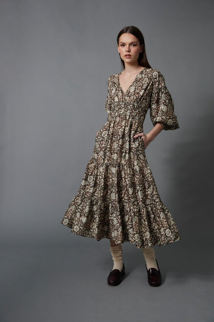 A woman stands against a gray background wearing the HOPE FOR FLOWERS TIERED PEASANT DRESS with puffed sleeves and a shirred bodice. She has her hands in its pockets and pairs dark loafers with light-colored socks.