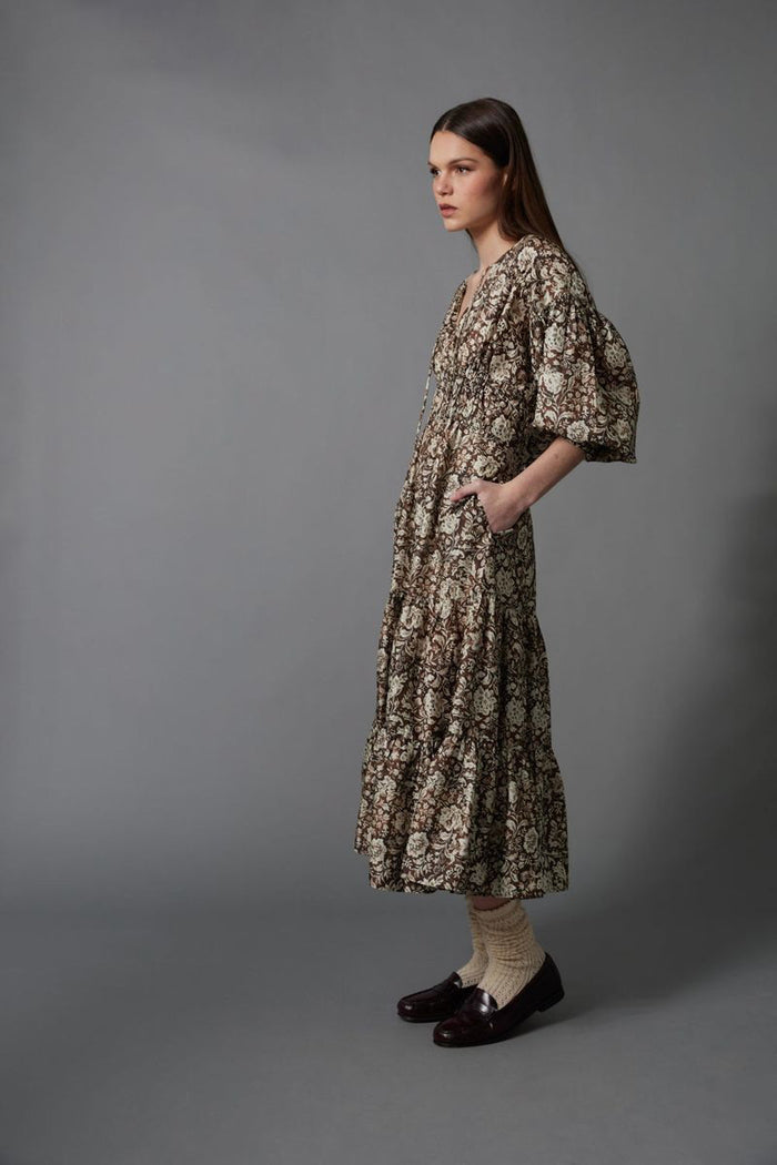 A woman stands against a gray background wearing the HOPE FOR FLOWERS TIERED PEASANT DRESS with puffed sleeves and a shirred bodice. She has her hands in its pockets and pairs dark loafers with light-colored socks.