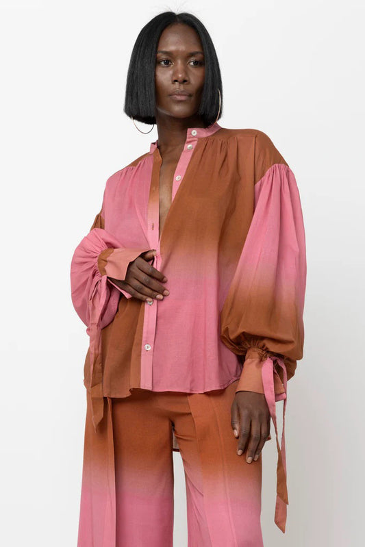 A person with straight black hair models the HOPE FOR FLOWERS WRIST TIE BLOUSE by HOPE FOR FLOWERS, featuring a flowy design in a pink and brown ombre, highlighted as sustainable clothing against a plain white background.