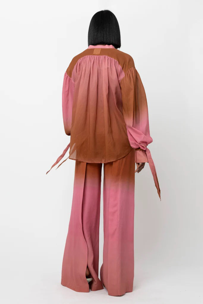 A person with straight black hair models the HOPE FOR FLOWERS WRIST TIE BLOUSE by HOPE FOR FLOWERS, featuring a flowy design in a pink and brown ombre, highlighted as sustainable clothing against a plain white background.