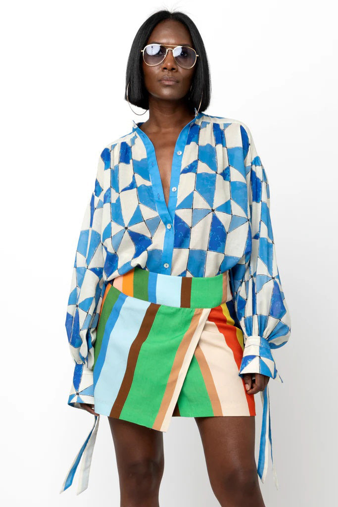 A woman in sunglasses and the HOPE FOR FLOWERS - WRIST TIE SHIRT by HOPE FOR FLOWERS, featuring a blue and white geometric pattern and long sleeves, stands confidently against a white background.