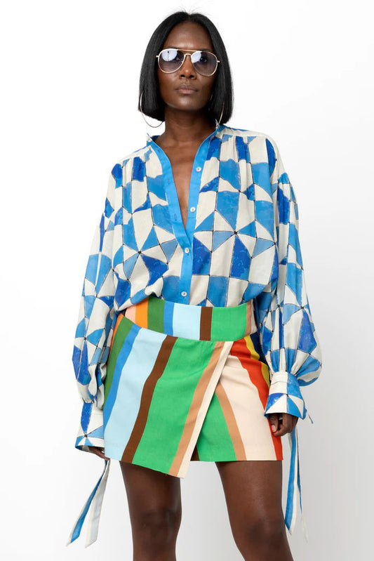 A woman in sunglasses and the HOPE FOR FLOWERS - WRIST TIE SHIRT by HOPE FOR FLOWERS, featuring a blue and white geometric pattern and long sleeves, stands confidently against a white background.