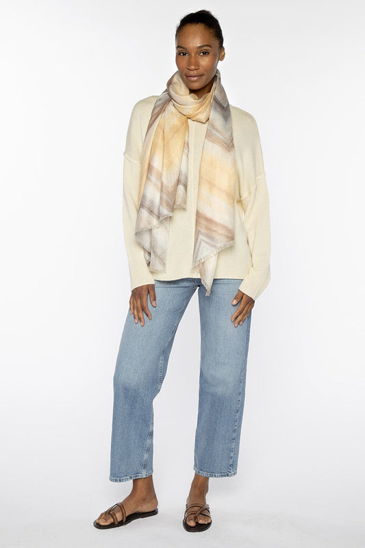 A woman stands against a white background wearing a cream sweater, light blue jeans, black sandals, and the KINROSS WAVE STRIPE SCARF IN CAMEO MULTI. She poses with one hand in her pocket, smiling softly.