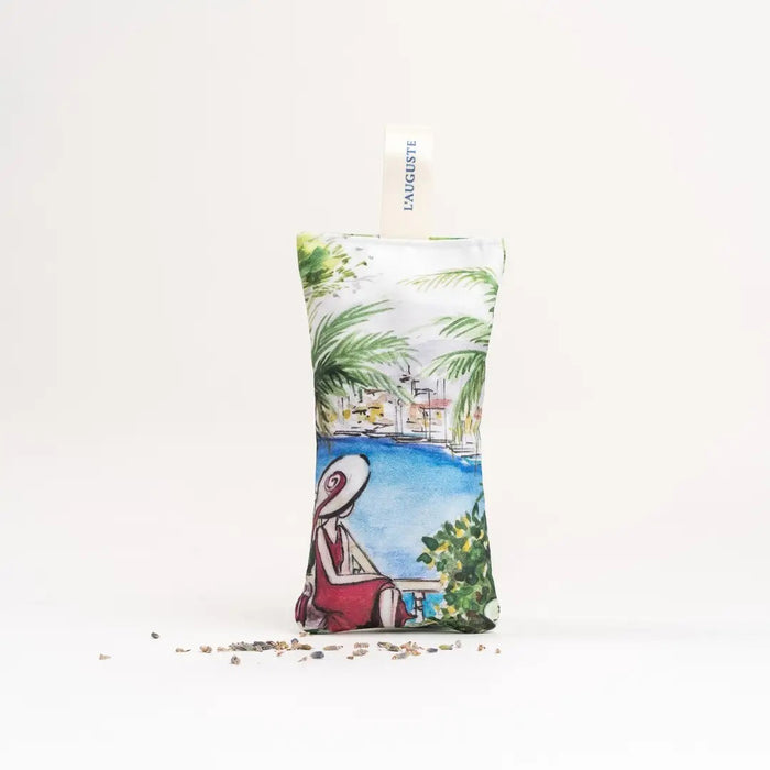 The L'AUGUSTE PROVENCE - FRENCH LAVENDER SACHET FROM PROVENCE is a small, handcrafted sachet featuring a hat-wearing woman by the water with palm trees, filled with organic lavender and labeled “L'AUGUSTE PROVENCE” for lasting freshness.