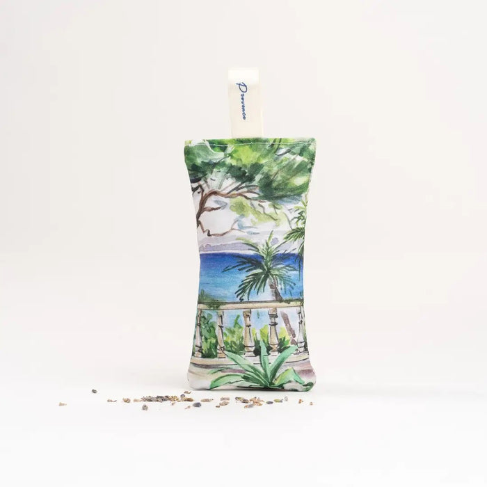 The L'AUGUSTE PROVENCE - FRENCH LAVENDER SACHET FROM PROVENCE is a small, handcrafted sachet featuring a hat-wearing woman by the water with palm trees, filled with organic lavender and labeled “L'AUGUSTE PROVENCE” for lasting freshness.
