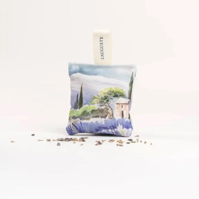 A sachet from L'AUGUSTE PROVENCE, featuring a watercolor landscape and "Provence" fabric tag, stands upright on a white surface amid dried lavender. This French artisanal gift comes in a FRENCH ORGANIC LAVENDER SACHET AND GIFT BOX.