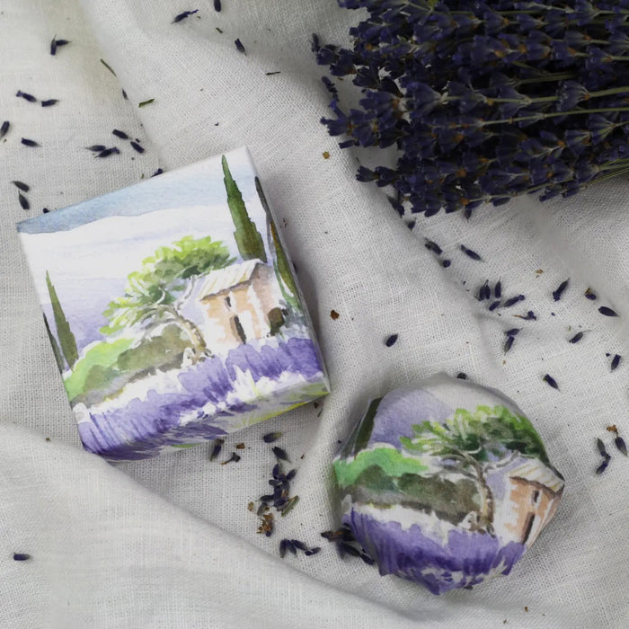 L'AUGUSTE PROVENCE's THE ESSENCE OF PROVENCE Lavender Soap with Gift Box, featuring handmade French soap and organic lavender soap in illustrated wrapping, is displayed on white fabric with dried lavender and a lavender bouquet nearby.