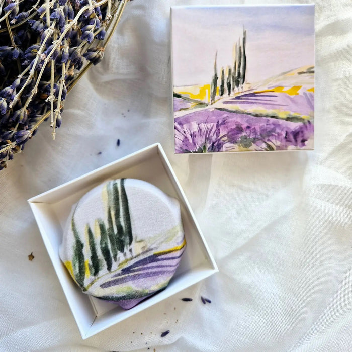 L'AUGUSTE PROVENCE - THE ESSENCE OF PROVENCE LAVENDER SOAP is a round, handmade French soap wrapped in painted landscape fabric, presented in a Provence gift box. Dried lavender rests nearby on a white cloth background.