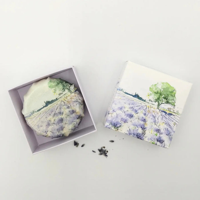 L'AUGUSTE PROVENCE – THE ESSENCE OF PROVENCE Lavender Soap, a round handmade French soap wrapped in cloth with a lavender field and tree motif, rests in an open L'AUGUSTE PROVENCE gift box beside its lid and scattered lavender buds.