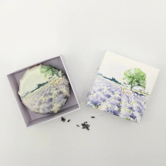 L'AUGUSTE PROVENCE – THE ESSENCE OF PROVENCE Lavender Soap, a round handmade French soap wrapped in cloth with a lavender field and tree motif, rests in an open L'AUGUSTE PROVENCE gift box beside its lid and scattered lavender buds.