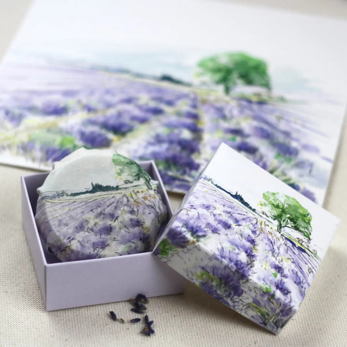 L'AUGUSTE PROVENCE – THE ESSENCE OF PROVENCE Lavender Soap, a round handmade French soap wrapped in cloth with a lavender field and tree motif, rests in an open L'AUGUSTE PROVENCE gift box beside its lid and scattered lavender buds.