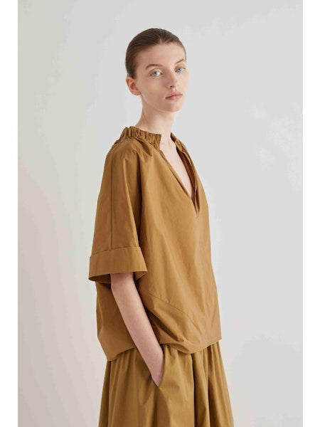 A woman with her hair in a bun stands in profile, wearing the MEIMEIJ Gathered Neck Blouse in Black by MEIMEIJ against a plain light background.