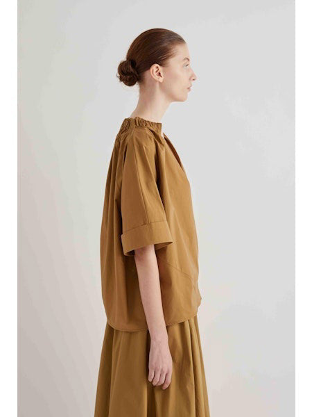 A woman with her hair in a bun stands in profile, wearing the MEIMEIJ Gathered Neck Blouse in Black by MEIMEIJ against a plain light background.