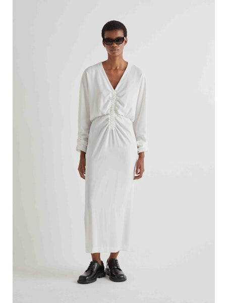 A person in profile wears the MEIMEIJ Gathered Waist Long Sleeve Dress in white, paired with black shoes and sunglasses. They have short hair and stand against a plain light background.