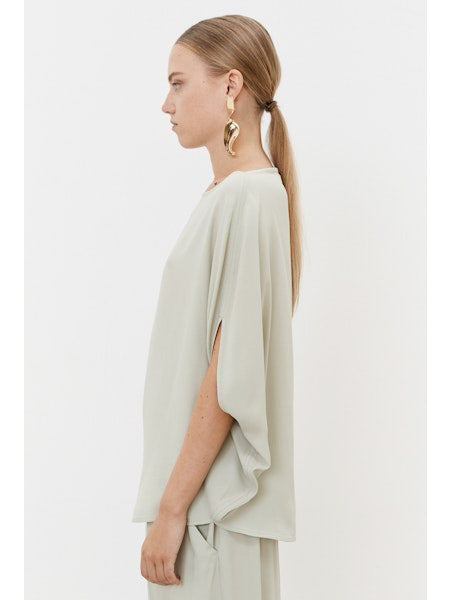 A person with tied-back hair faces away, wearing a light gray MEIMEIJ Silk Blend Top with minimalist design and elegant acetate-silk earrings against a plain white background.
