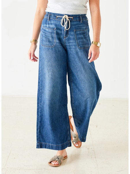 Wearing MERSEA's LONDONBERRY Drawstring Supersoft Denim Pant with wide legs and large front pockets, paired with a light grey top, gold jewelry, and brown sandals, a person poses against a plain light background.